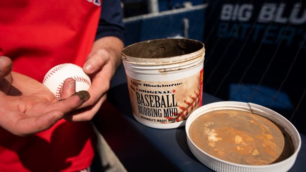 Person in a red sox uniform holds a baseball next to a container of rubbing mud