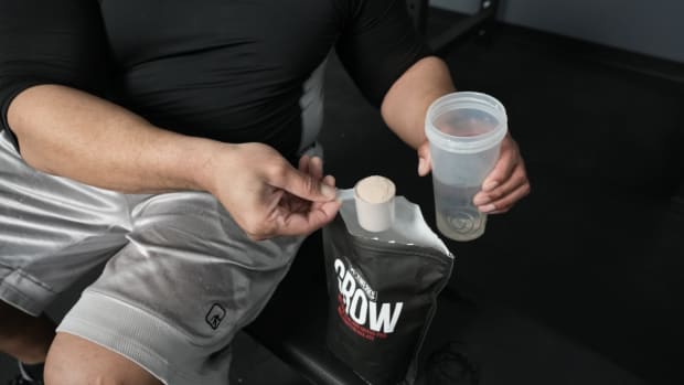 A man sitting on a weight bench in a gym and pouring XWerks Grow whey protein powder into a shaker bottle