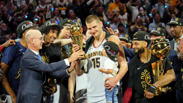 Nuggets center Nikola Jokic holds the 2023 NBA Finals MVP award while the team celebrates the title.