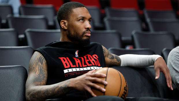 Former Trail Blazers guard Damian Lillard stares while sitting on the sideline holding a basketball.
