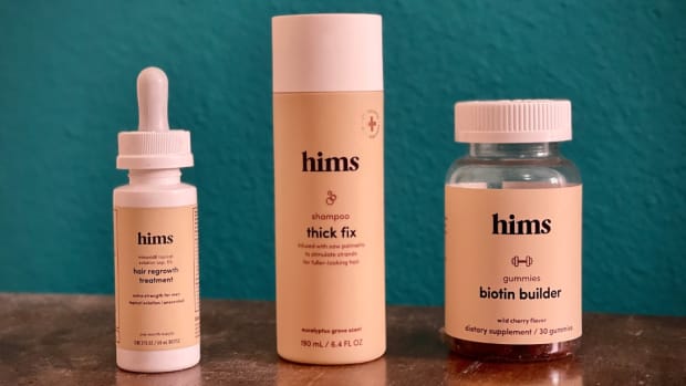 The Hims hair growth serum, shampoo and biotin builder gummies on a wooden table with a blue background.