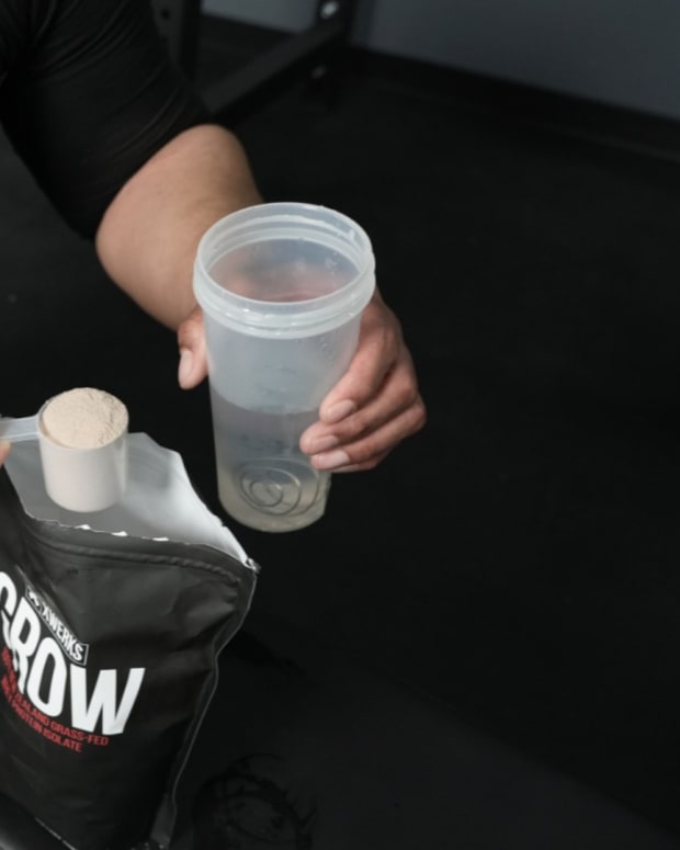 A man sitting on a weight bench in a gym and pouring XWerks Grow whey protein powder into a shaker bottle