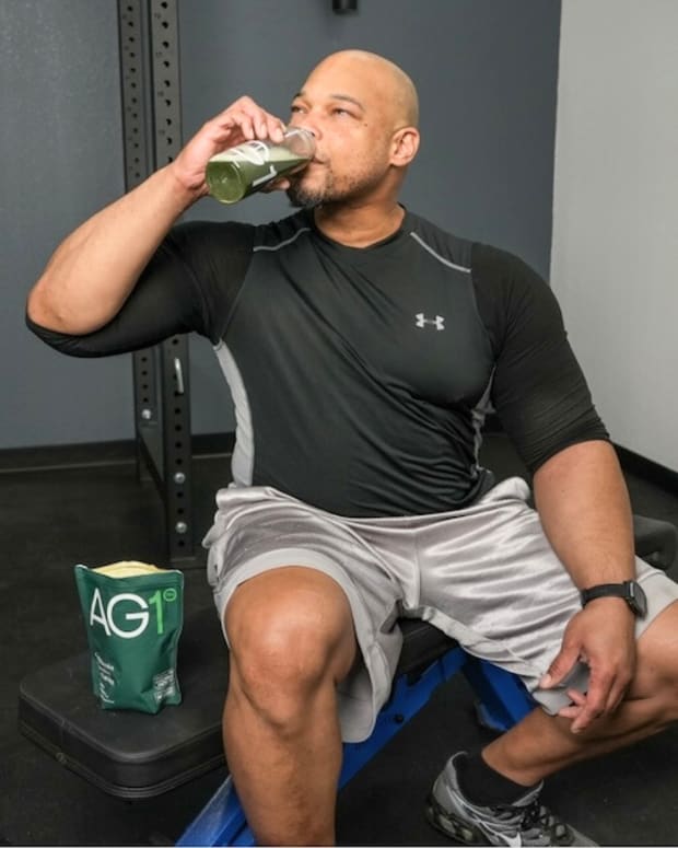 A man dressed in workout clothes drinks AG1 greens powder from a shaker bottle.
