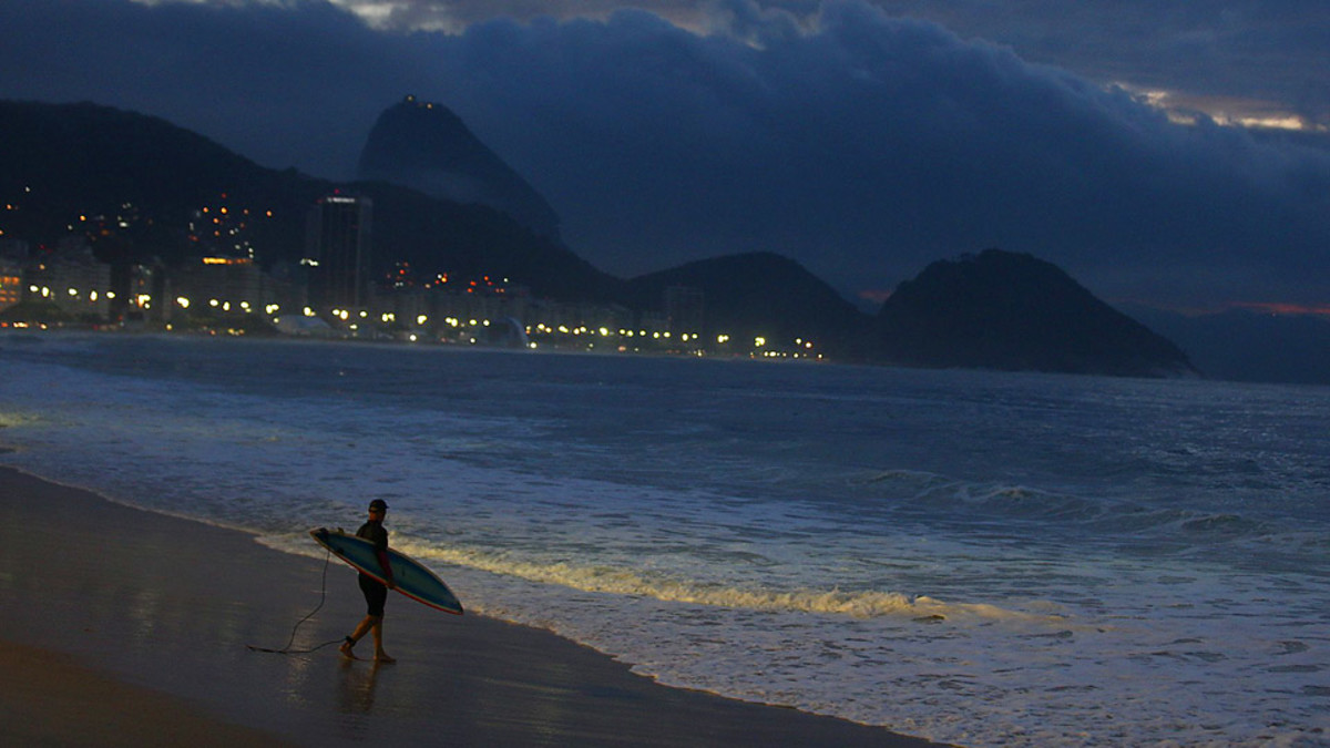 At nearly sunrise in the second week of the World Cup, a lone surfer walks toward the waves at a mostly empty Copacabana Beach. 