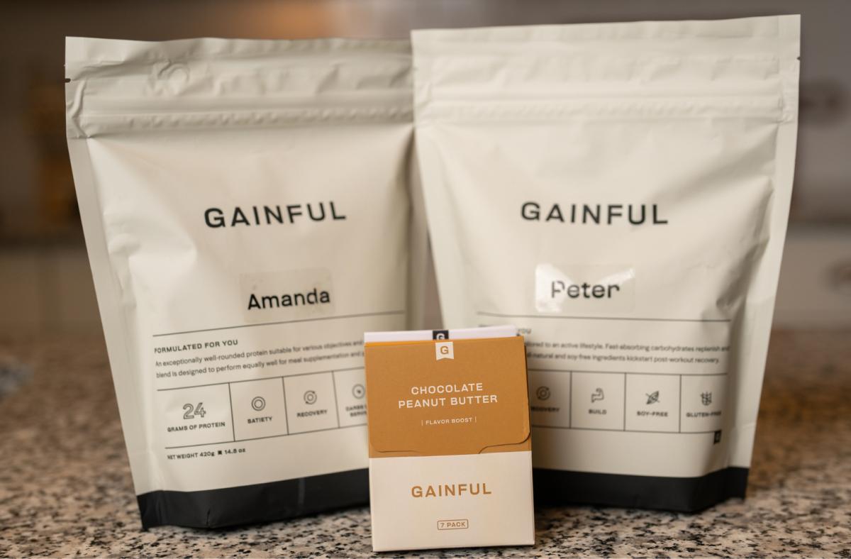 2 Gainful protein powder white bags one labeled "Amanda" and the other labeled "Peter" with a small chocolate peanut butter sample in front sitting on a counter top