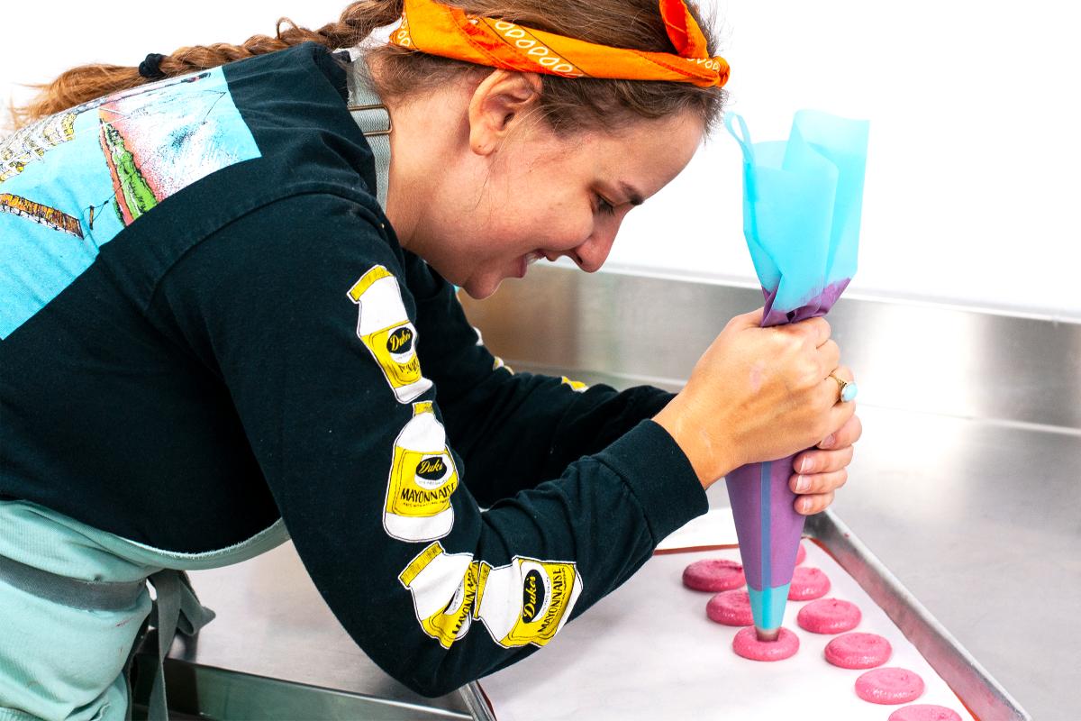 A woman pipes macarons onto a sheet wearing a black long sleeve with mayo jar decals on it