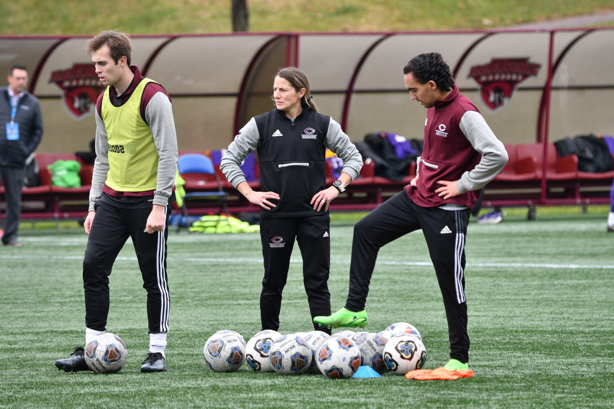 Coach Julianne Sitch stands in the middle of two players, one of whom is about to dribble the soccer ball