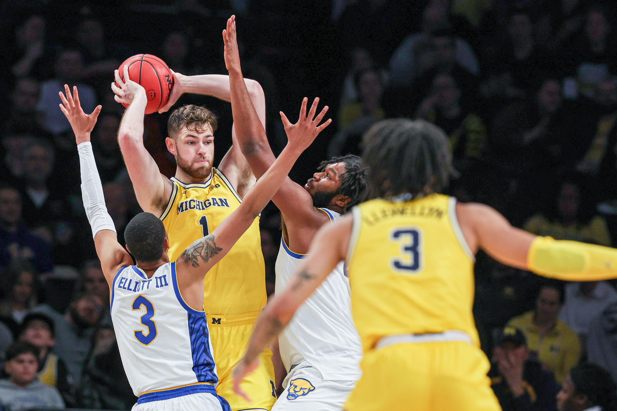 Michigan player tries to make a pass around two defenders in college hoops game.