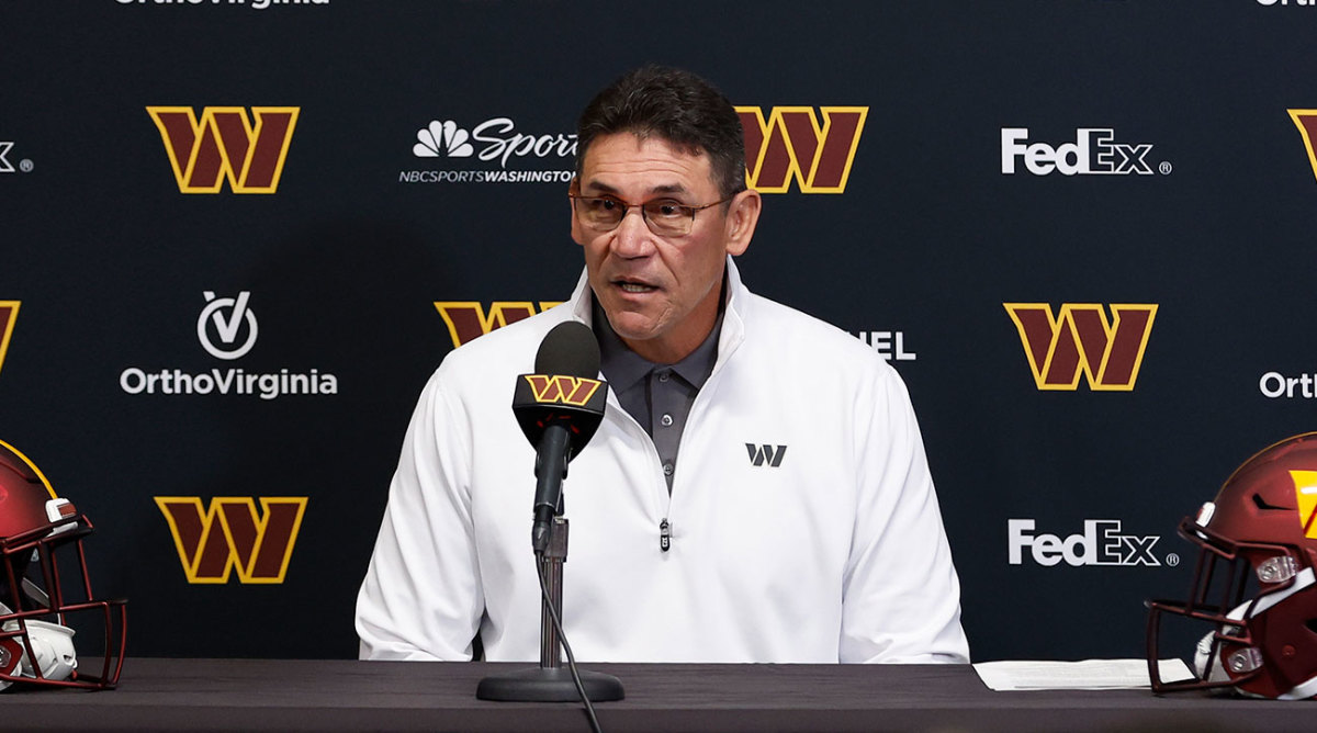 Ron Rivera sits at a podium in front of a Commanders backdrop