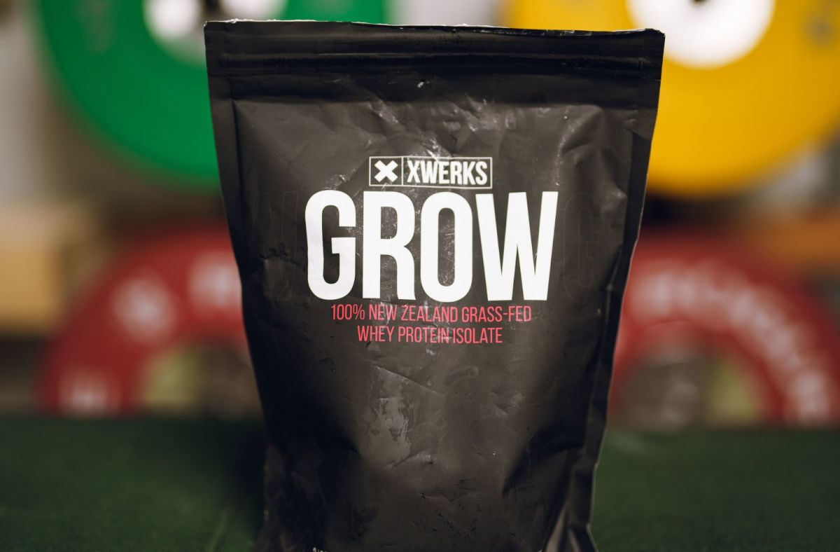 A black bag of XWerks Grow whey protein isolate against background of colorful weight plates