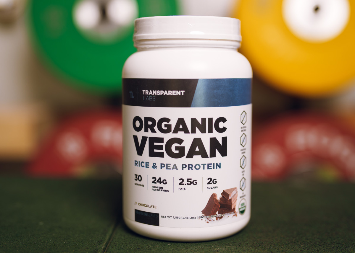 Transparent Labs Organic Vegan Rice and Pea Protein chocolate flavor in white canister with blue and black label on colorful background of blurred weight plates