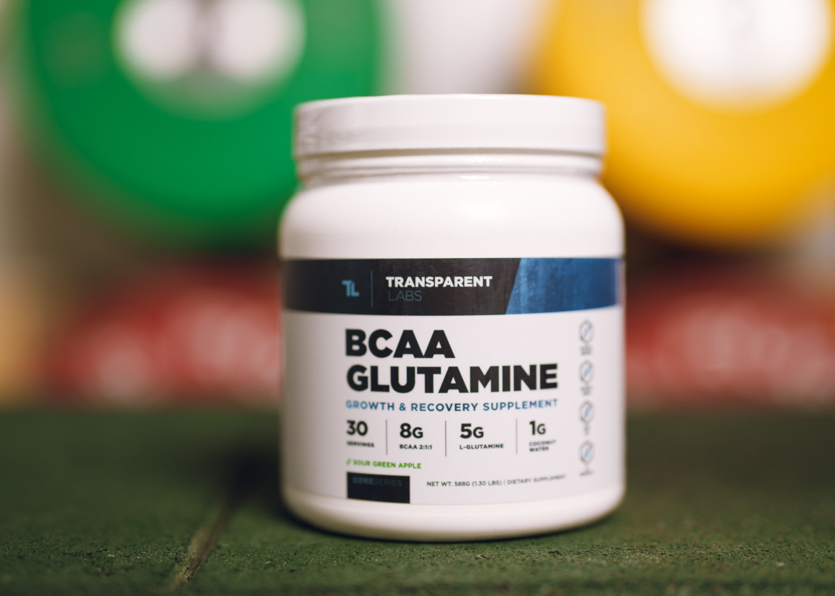 Transparent Labs BCAA Glutamine in white canister, label with blue and black text on a colorful background of blurred weight plates