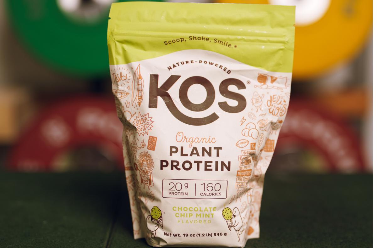 A bag of KOS Organic Plant Protein in Chocolate Chip Mint flavor and blurred weight plates in the background