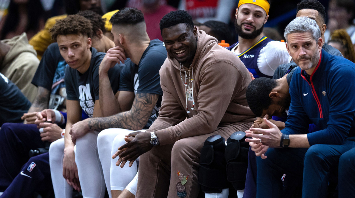 New Orleans Pelicans forward Zion Williamson, dressed in street clothes, laughs on the bench with forward Garrett Temple.