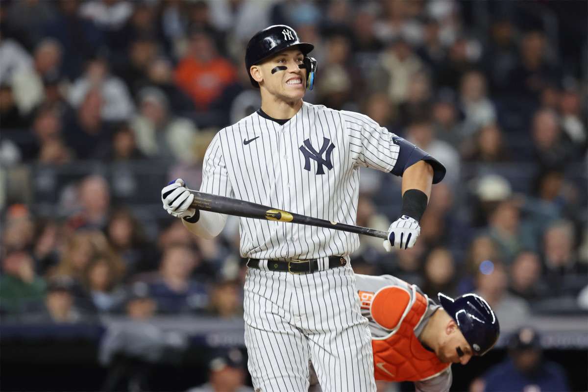 Yankees star slugger Aaron Judge stands on the field holding a bat and smiling