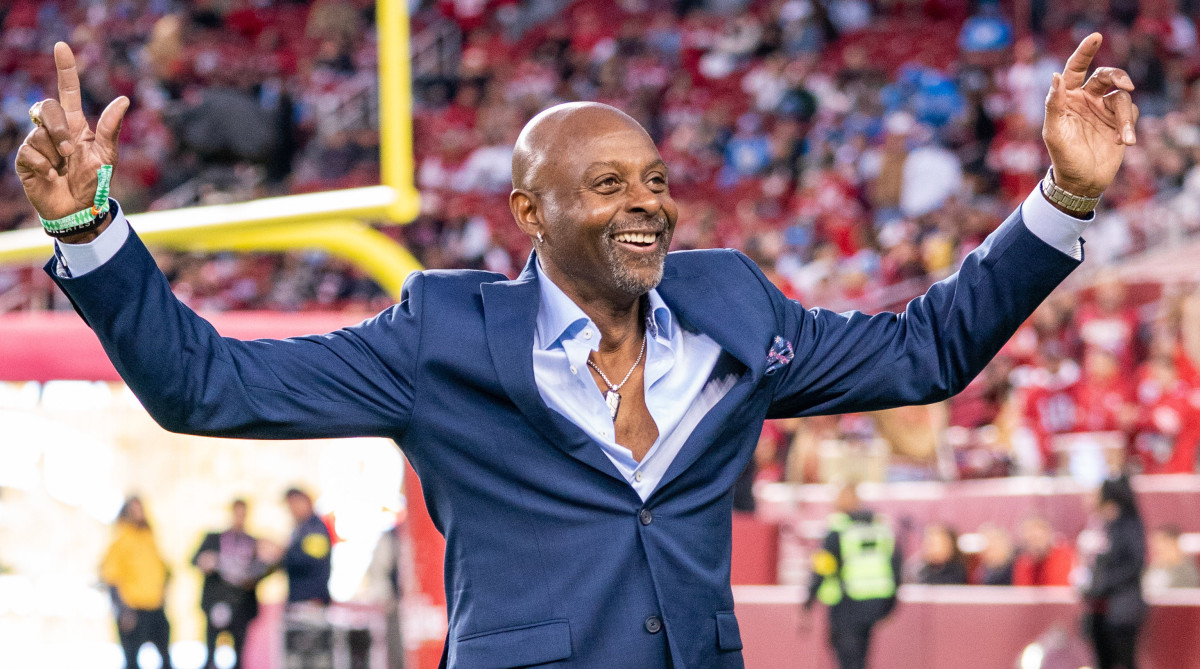 Former 49ers receiver Jerry Rice, wearing a suit, holds his hands up on the field before a game