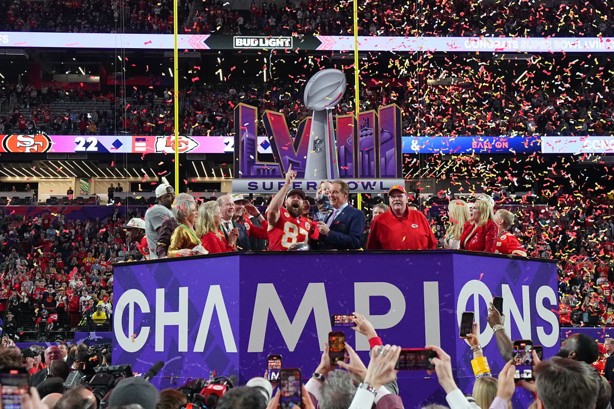 Travis Kelce points up while standing with Andy Reid and other Chiefs members in front of a Super Bowl LVIII sign and on top of a “Champions” platform