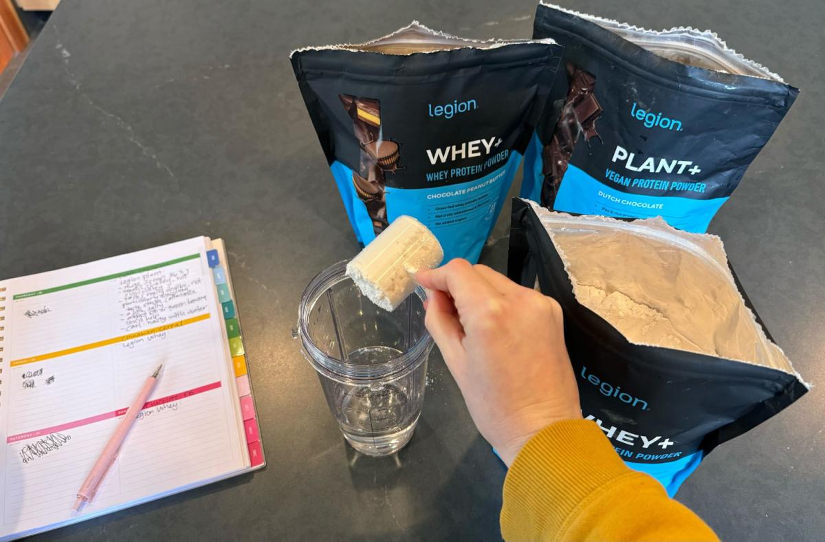 A person adding a scoop of Legion Whey+ protein powder in Cinnamon Cereal flavor to a blender bottle of water next to a notepad with testing notes