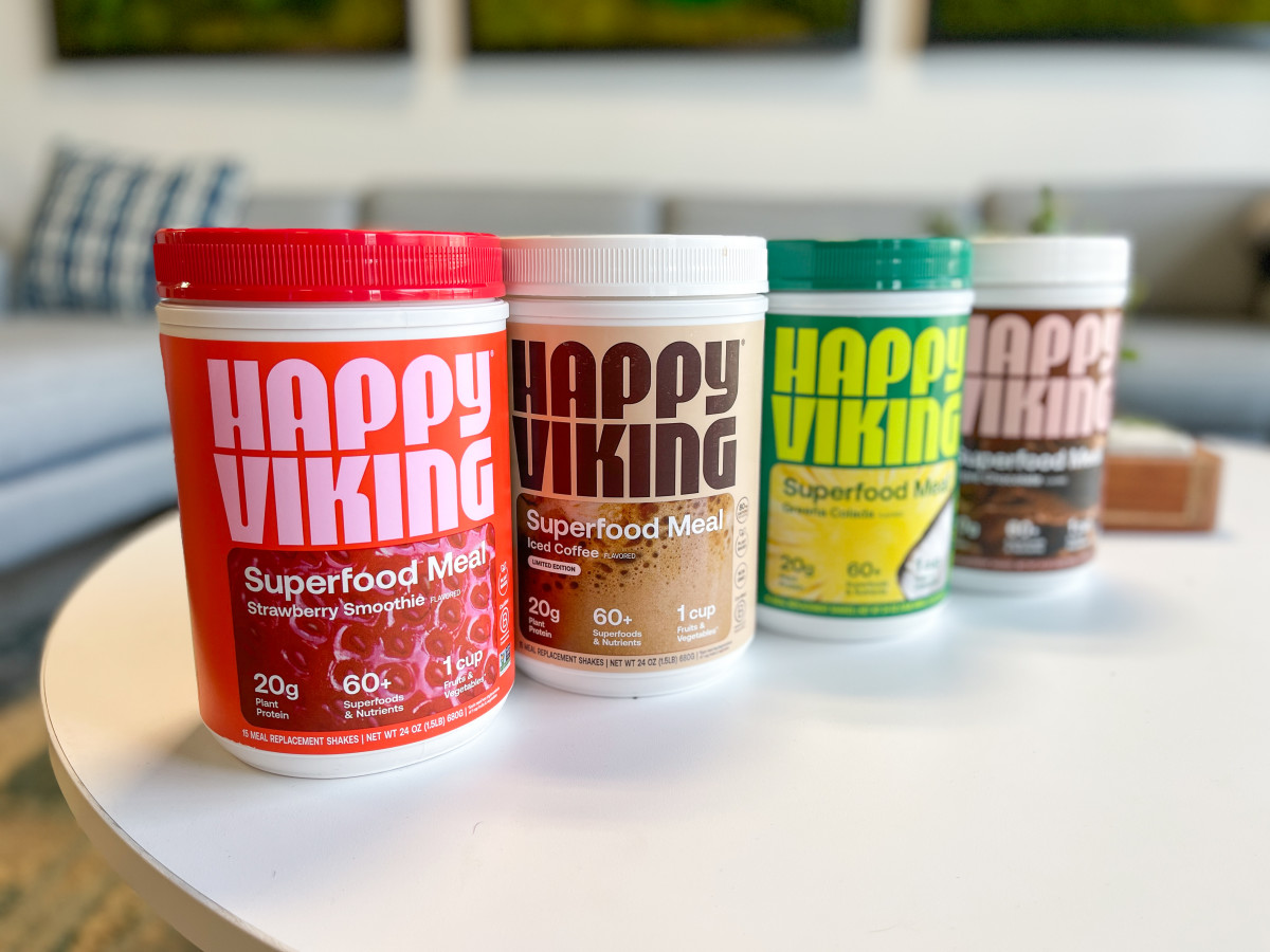 Four containers of Happy Viking Superfood Meal protein powder on a white table in flavors Strawberry Smoothie, Iced Coffee, Greena Colada, and Triple Chocolate