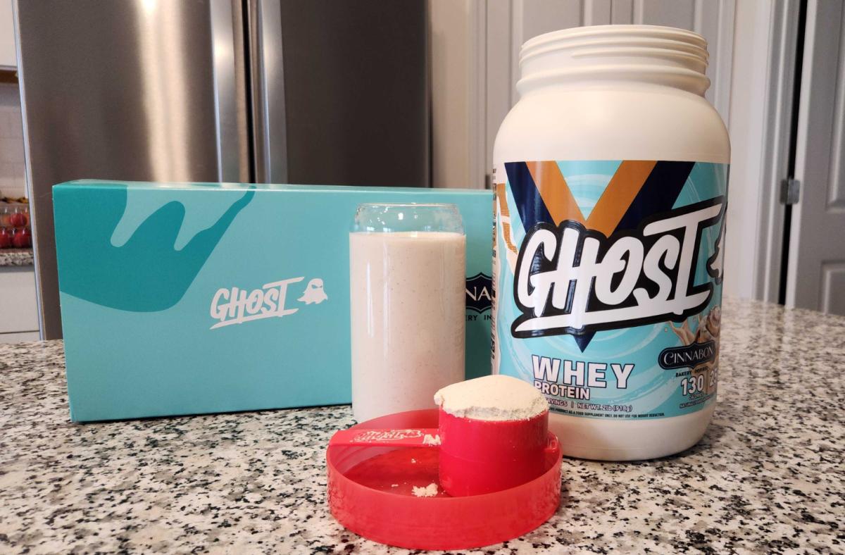 A container of Ghost Whey Protein powder in Cinnabon flavor next to a glass of blended Ghost Whey protein and a scoop full of Ghost Whey protein powder on a granite countertop