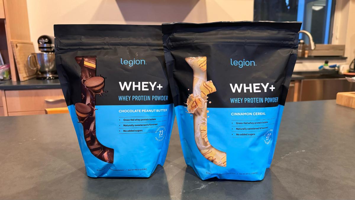 Two bags of Legion Whey+ protein powder in Chocolate Peanut Butter and Cinnamon Cereal flavors on a kitchen counter