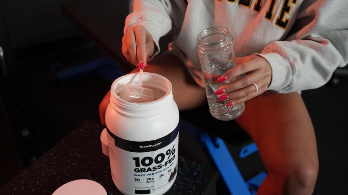 A woman in a gym setting scooping Transparent Labs whey protein powder into a shaker bottle full of water