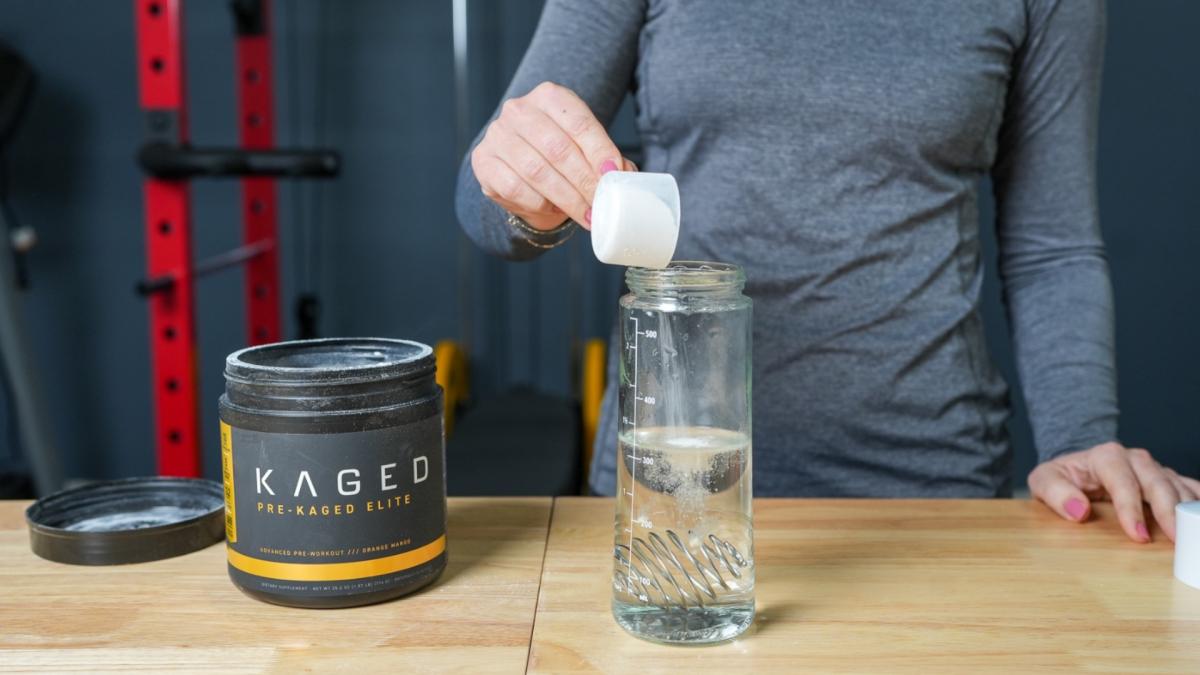 A woman in a gym scooping Kaged Pre-Kaged Elite pre-workout powder into a shaker bottle half full of water
