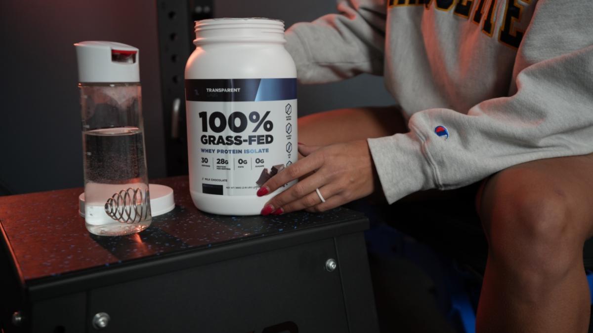 A woman in a gym preparing to scoop Transparent Labs whey protein isolate powder into a shaker bottle filled with water