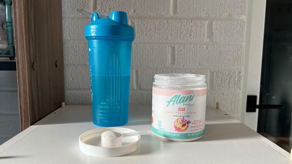 A container of Alani Nu BCCA powder in Sour Gummy flavor and a shaker bottle full of water on a kitchen counter