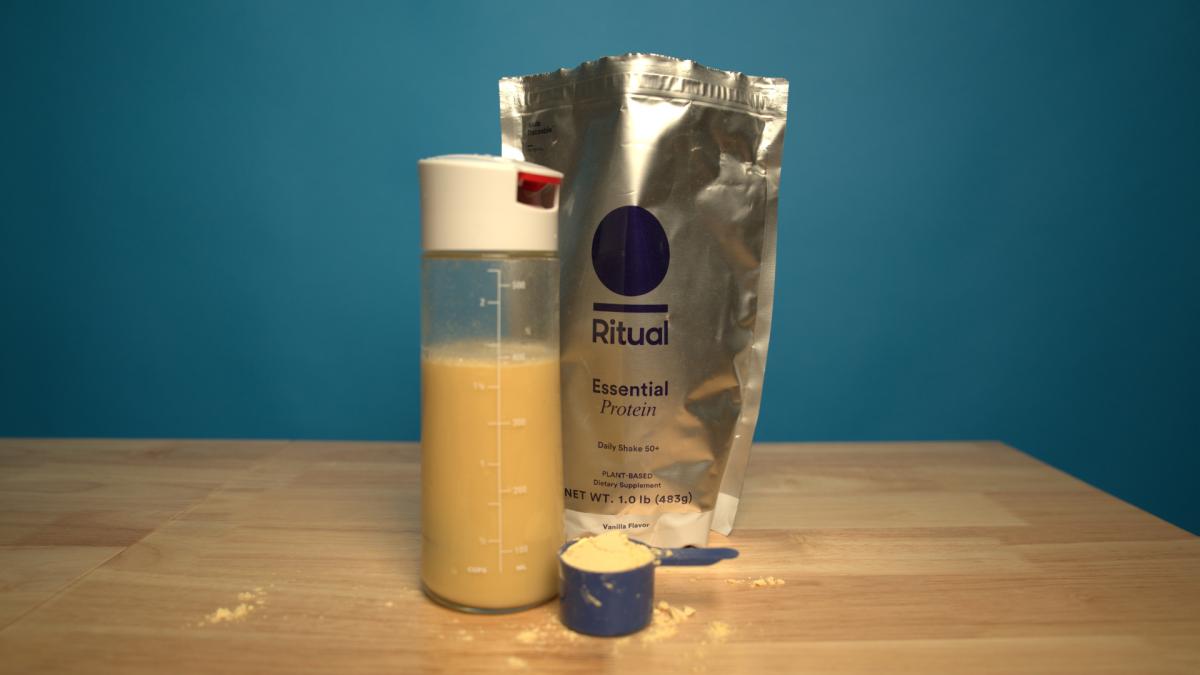 A bag of Ritual Essential Protein 50+ and a shaker bottle with Ritual protein mixed with water on a wooden table with a blue backdrop