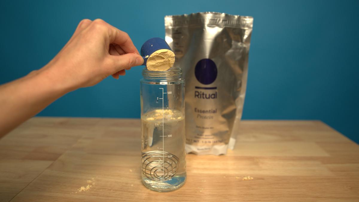 A person scooping Ritual Essential 50+ protein powder into a clear shaker bottle filled with water