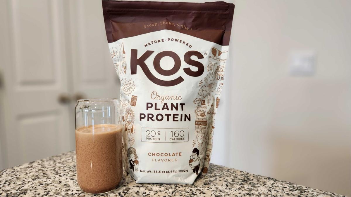 A bag of KOS plant protein in Chocolate flavor and a protein shake in a glass cup on a granite counter