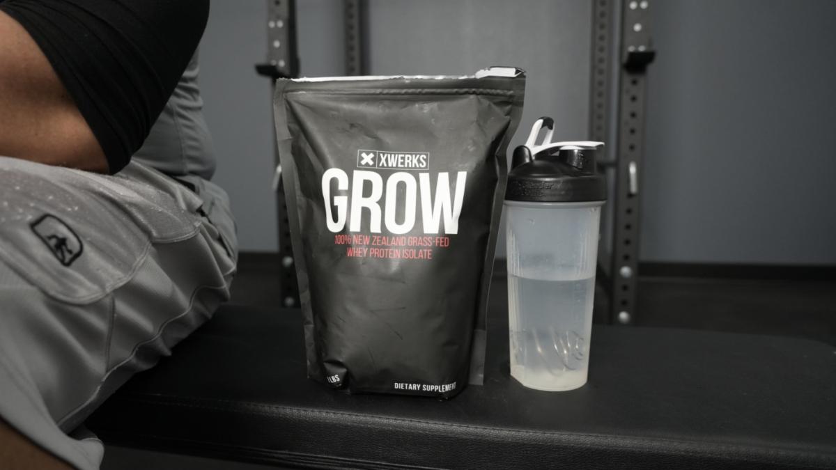 The front side of a bag of XWerks Grow whey protein powder and a shaker bottle with water on a weight bench in a gym