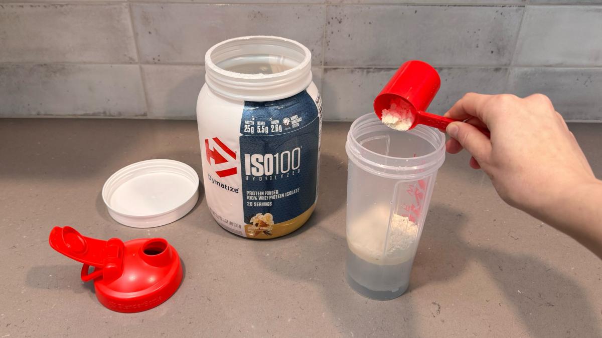 A person scooping Dymatize ISO100 Hydrolyzed Whey Protein Powder in Gourmet Vanilla flavor into a shaker bottle with water