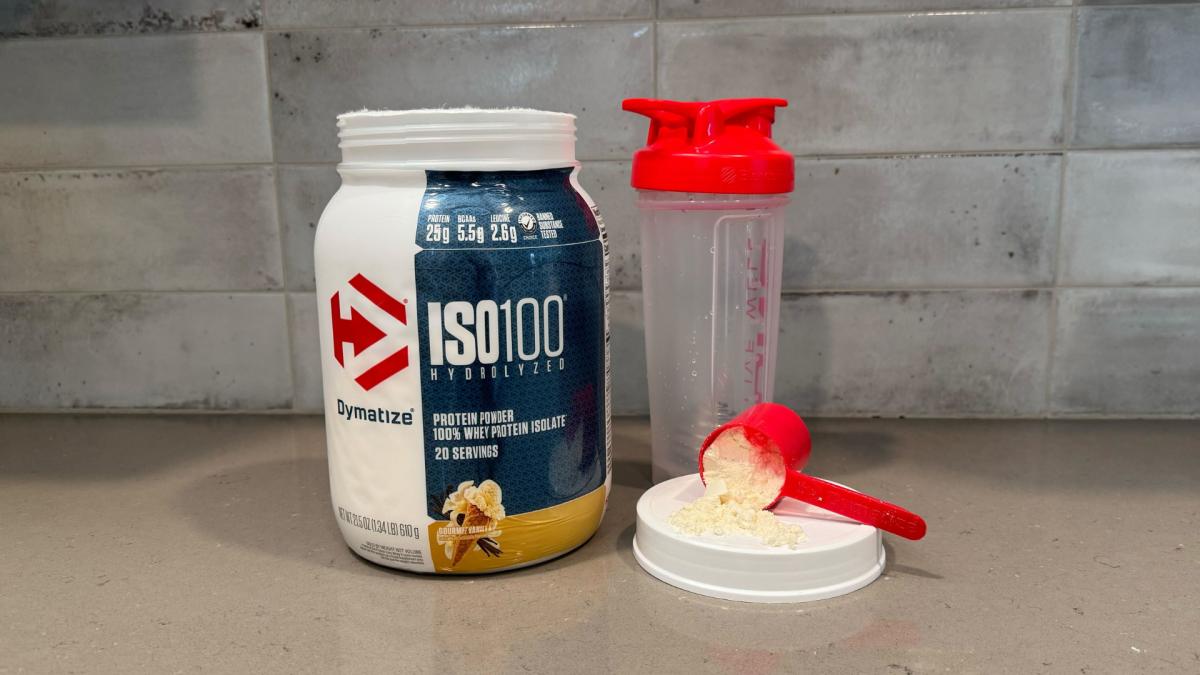A kitchen counter with a container of Dymatize ISO100 Hydrolyzed Whey Protein Powder in Gourmet Vanilla flavor, a red scoop of Dymatize protein powder and a shaker bottle