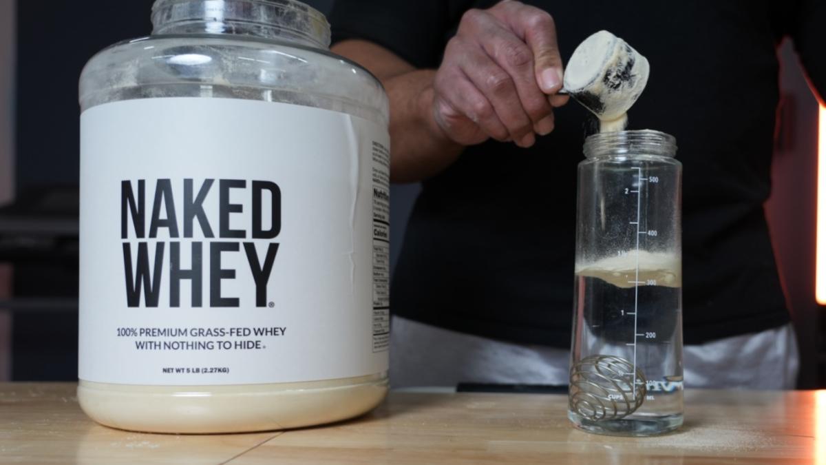A man pouring a scoop of unflavored Naked Whey protein powder into a clear shaker bottle half filled with water