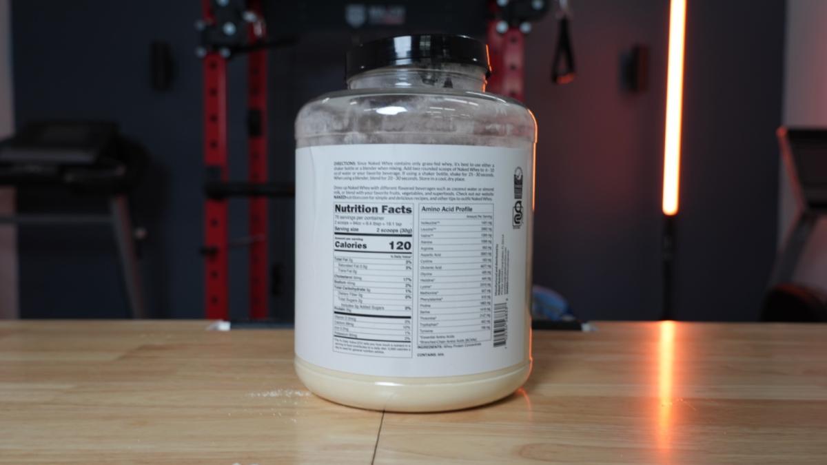 The backside of a five-pound tub of unflavored Naked Whey protein powder showing the Nutrition Facts with gym equipment in the background