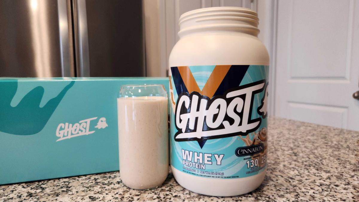 A tub of Ghost Whey Protein in Cinnabon flavor next to a Ghost Whey protein shake in a clear glass on a kitchen counter