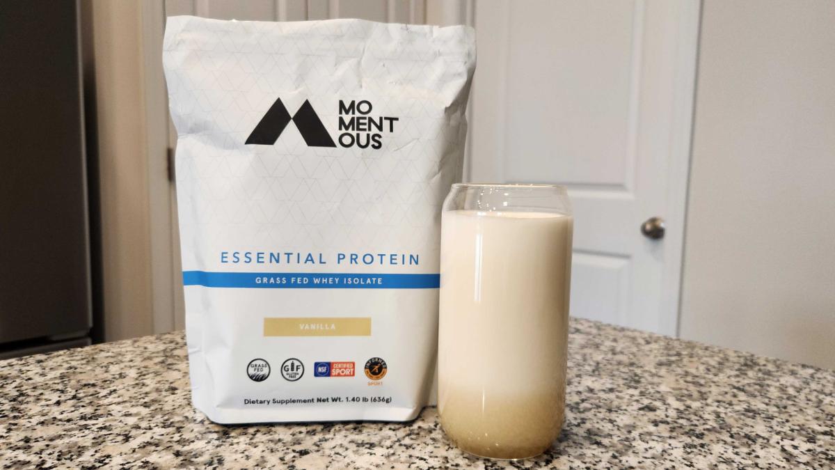 A bag of Momentous Essential Protein Grass-Fed Whey Isolate in Vanilla flavor and a glass of Momentous whey mixed with water on a granite kitchen counter