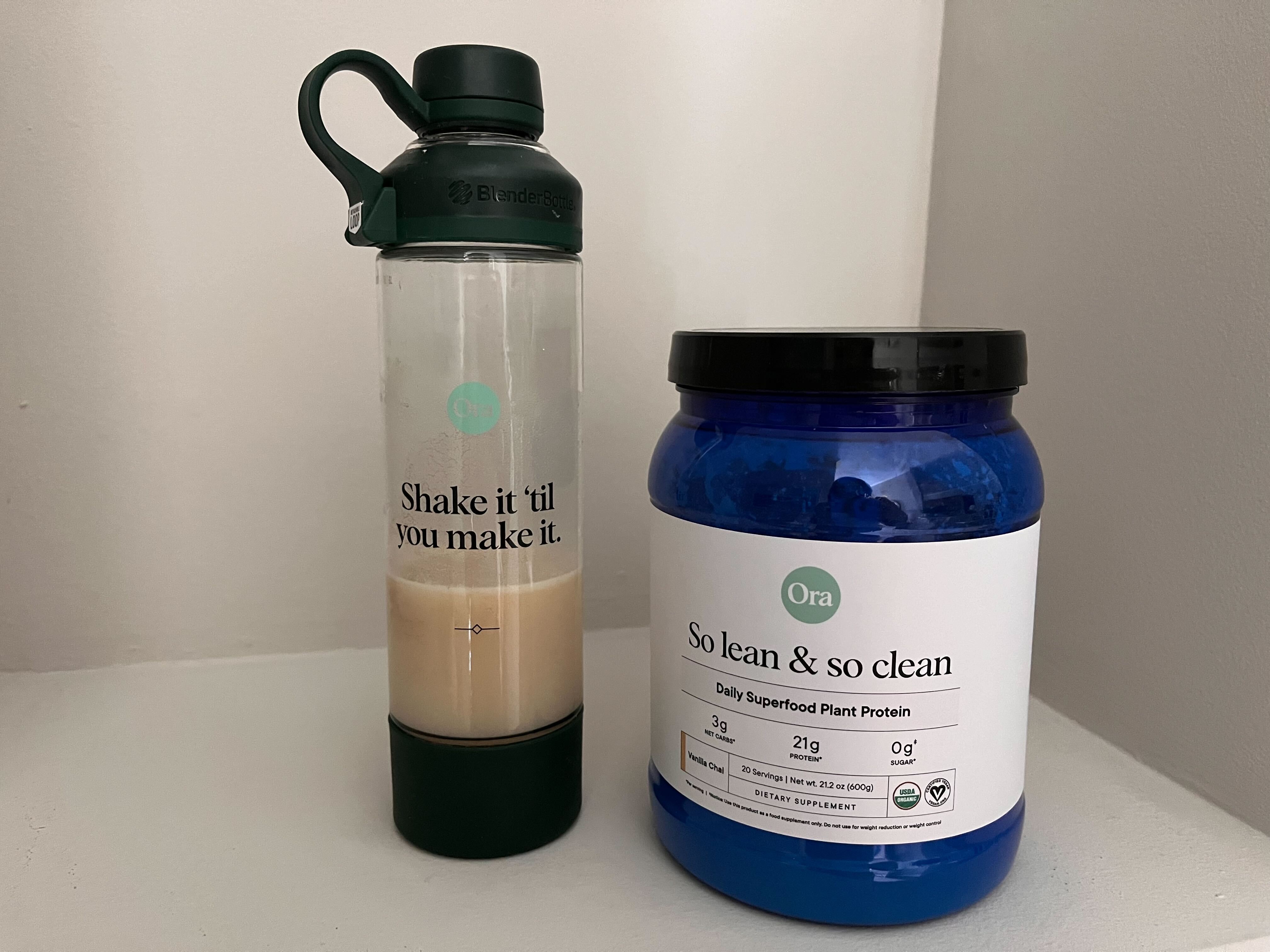 A tub of Ora plant protein powder on a white table next to a shaker bottle with mixed Ora protein powder and water