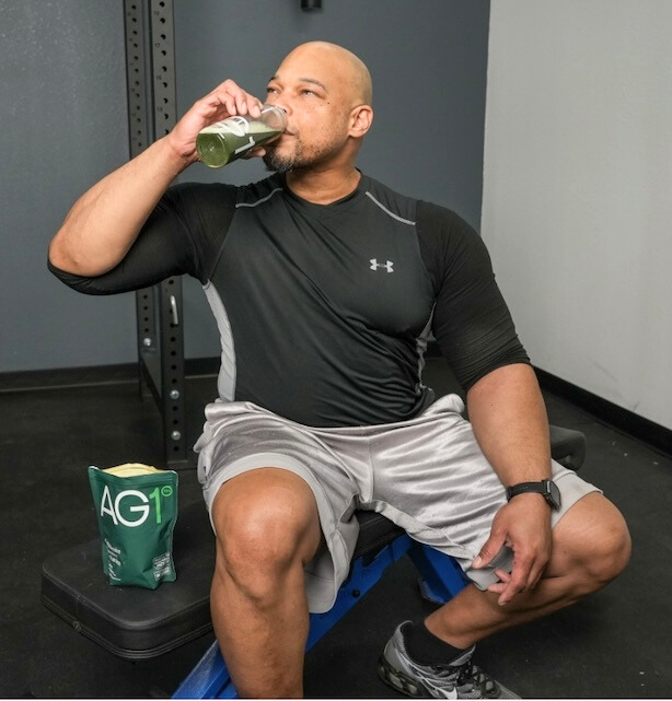 A man dressed in workout clothes drinks AG1 greens powder from a shaker bottle.