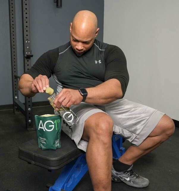 A man dressed in workout clothes puts a scoop of AG1 greens powder into a mixer bottle filled halfway with water.