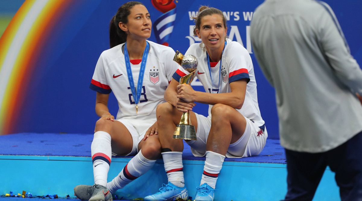 Tobin Heath holds the Women's World Cup trophy while sitting next to Christen Press after the USWNT won in 2019.