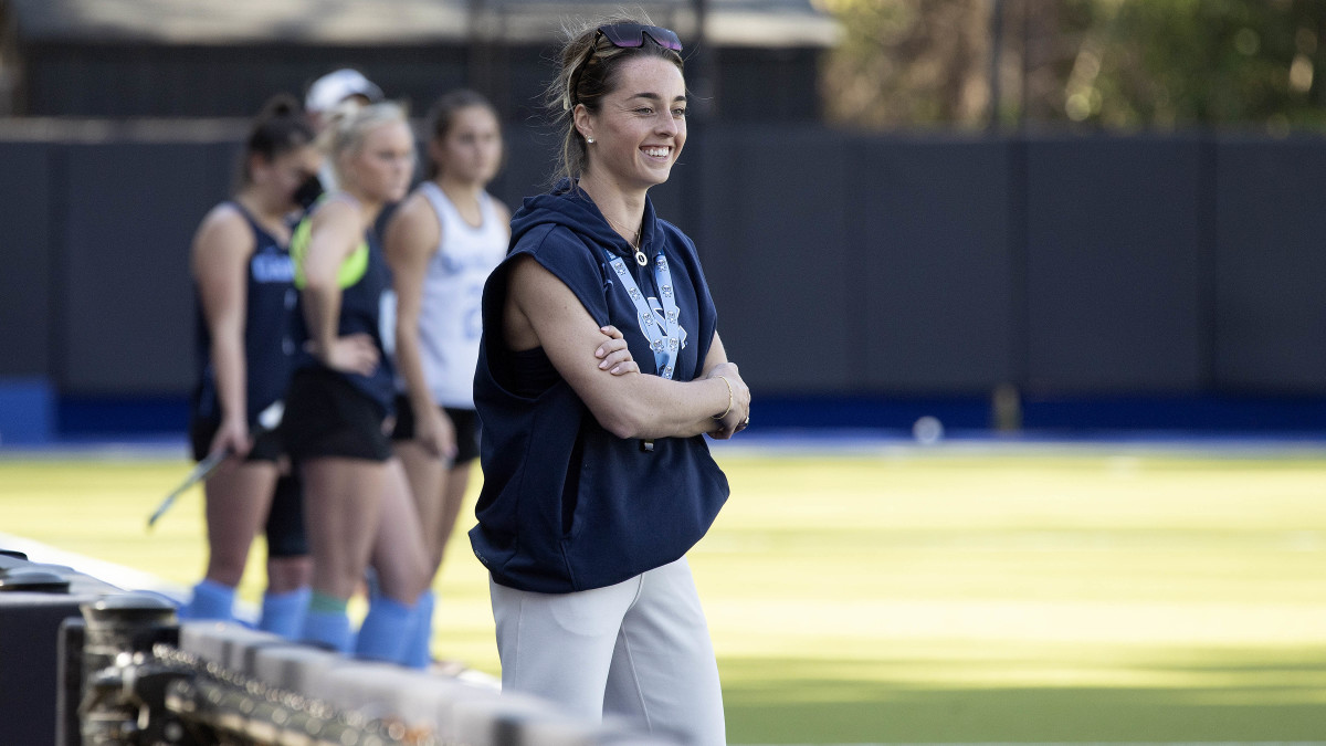 UNC field hockey coach Erin Matson smiles while watching practice with players standing behind her.