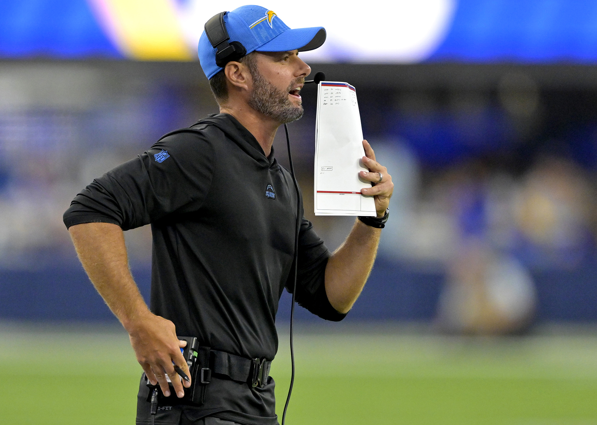 Brandon Staley talks into a headset on the sideline while holding a piece of paper up to cover his mouth