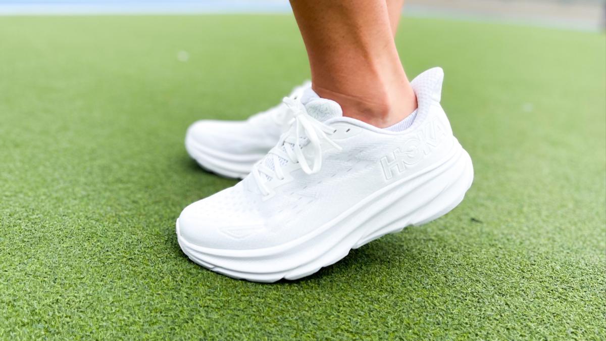 Hoka Clifton 9 shoes in white on shoe model standing on turf.