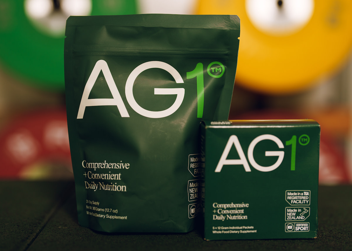 AG1 greens powder in pouch size and travel packs in a small box with blurred weight plates in the background