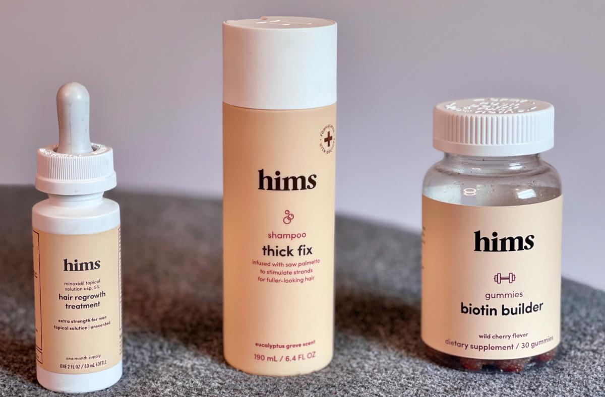A bottle of hims hair regrowth treatment, hims thick fix shampoo and hims biotin builder gummies against a grey background.