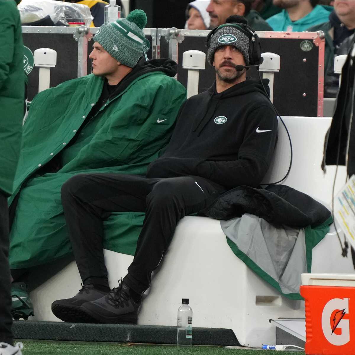 Aaron Rodgers sits on the bench wearing a headset next to Zach Wilson, who is wrapped in a green coat