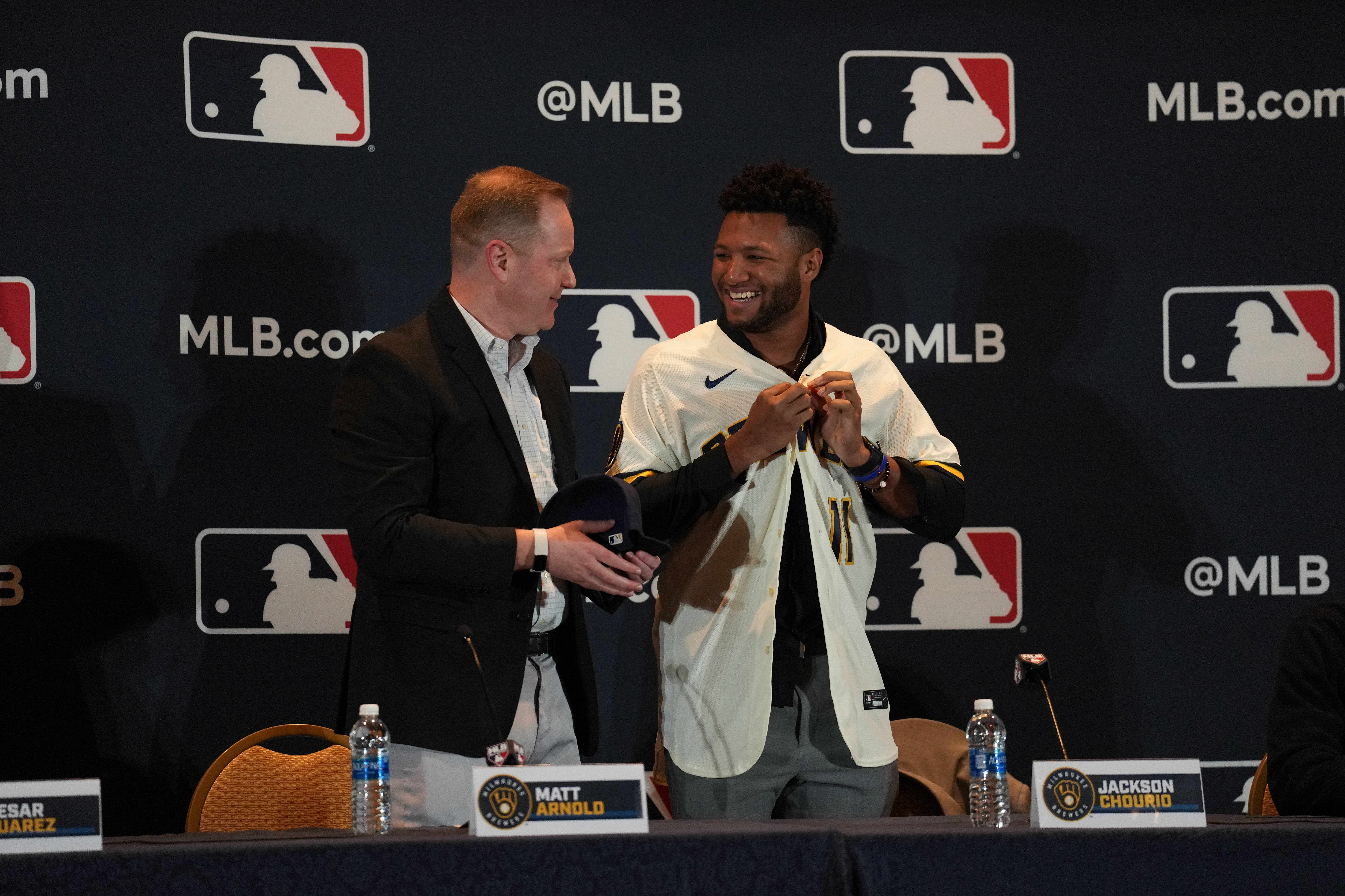 Jackson Chourio puts on a Brewers jersey smiling at GM Matt Arnold while the two stand in front of a MLB backdrop